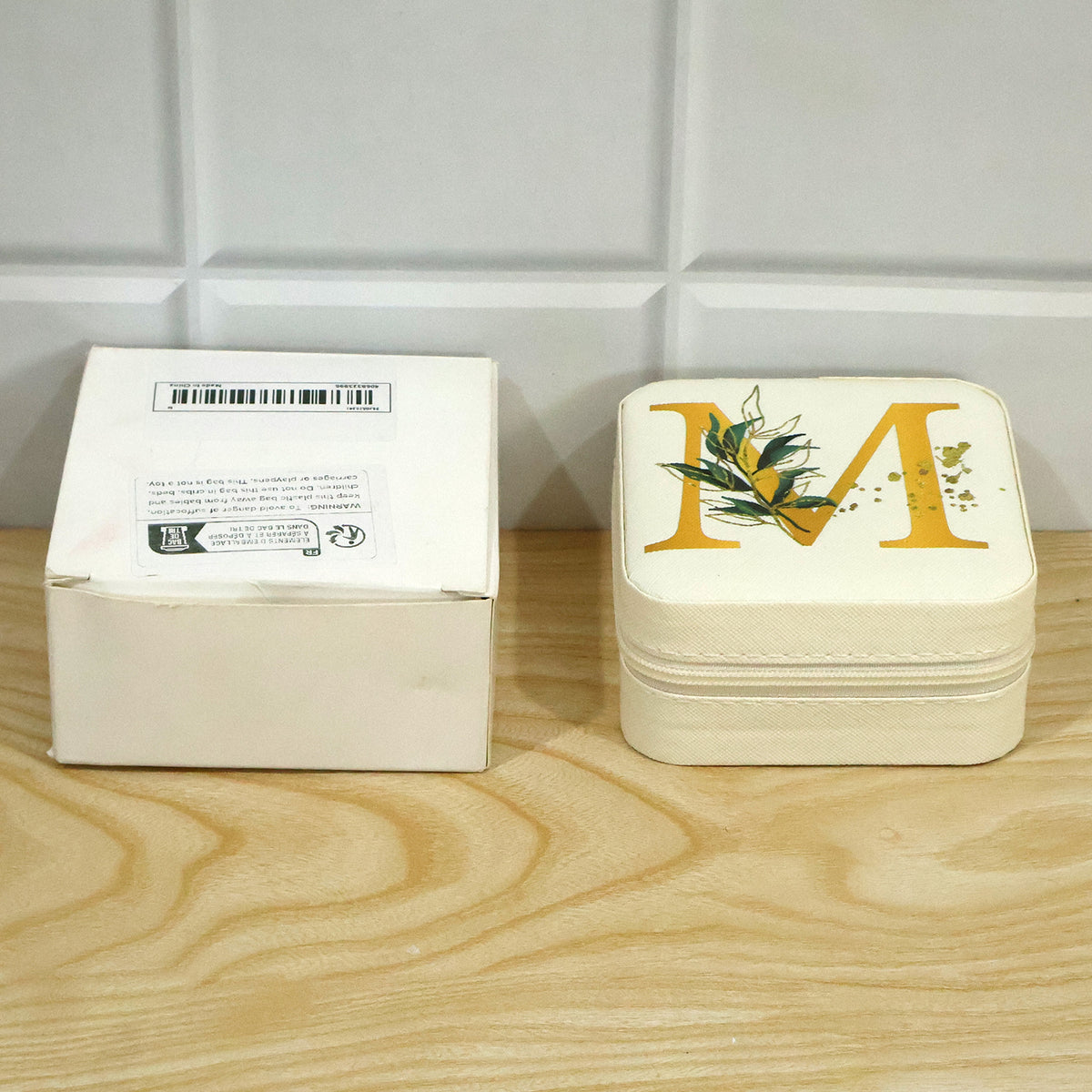 A closed beige jewellery box with a gold 'M' sits beside a plain white cardboard box on a light wooden surface.