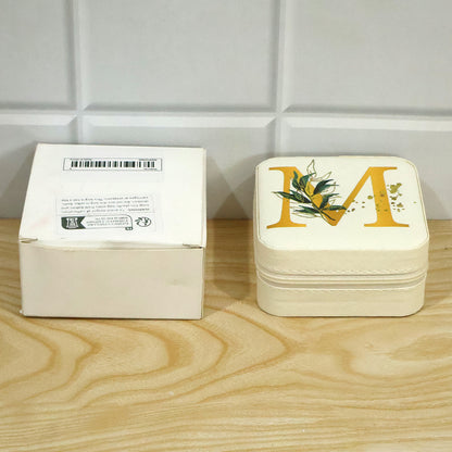 A closed beige jewellery box with a gold 'M' sits beside a plain white cardboard box on a light wooden surface.
