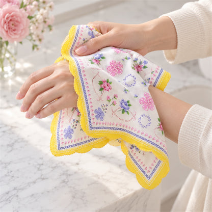 Two hands gently hold a white floral handkerchief with a yellow scalloped edge over a marble surface, showing its soft