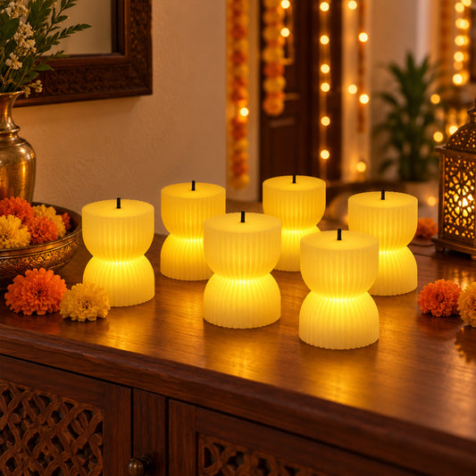 Six hourglass-shaped ribbed LED candles glowing warmly on a wooden console table with orange and yellow flowers.