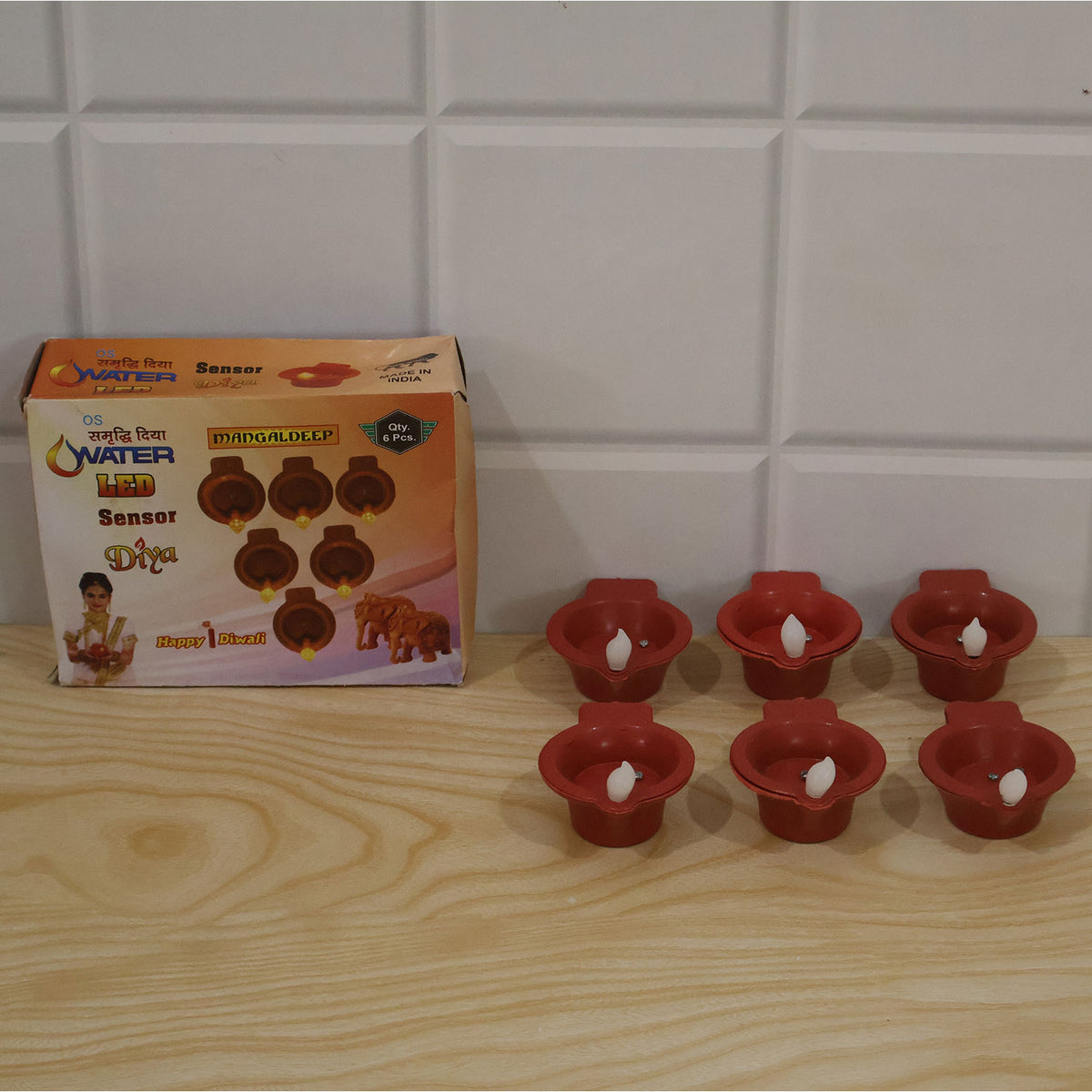 Six red plastic diyas with white LED flames, next to their product box, on a light wooden surface against a tiled wall.