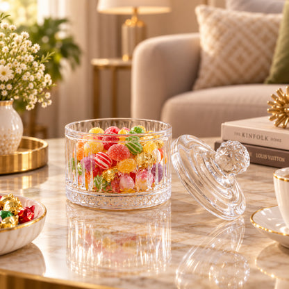 Open clear glass candy bowl with its lid resting beside it, revealing the bright candies inside on a reflective table.