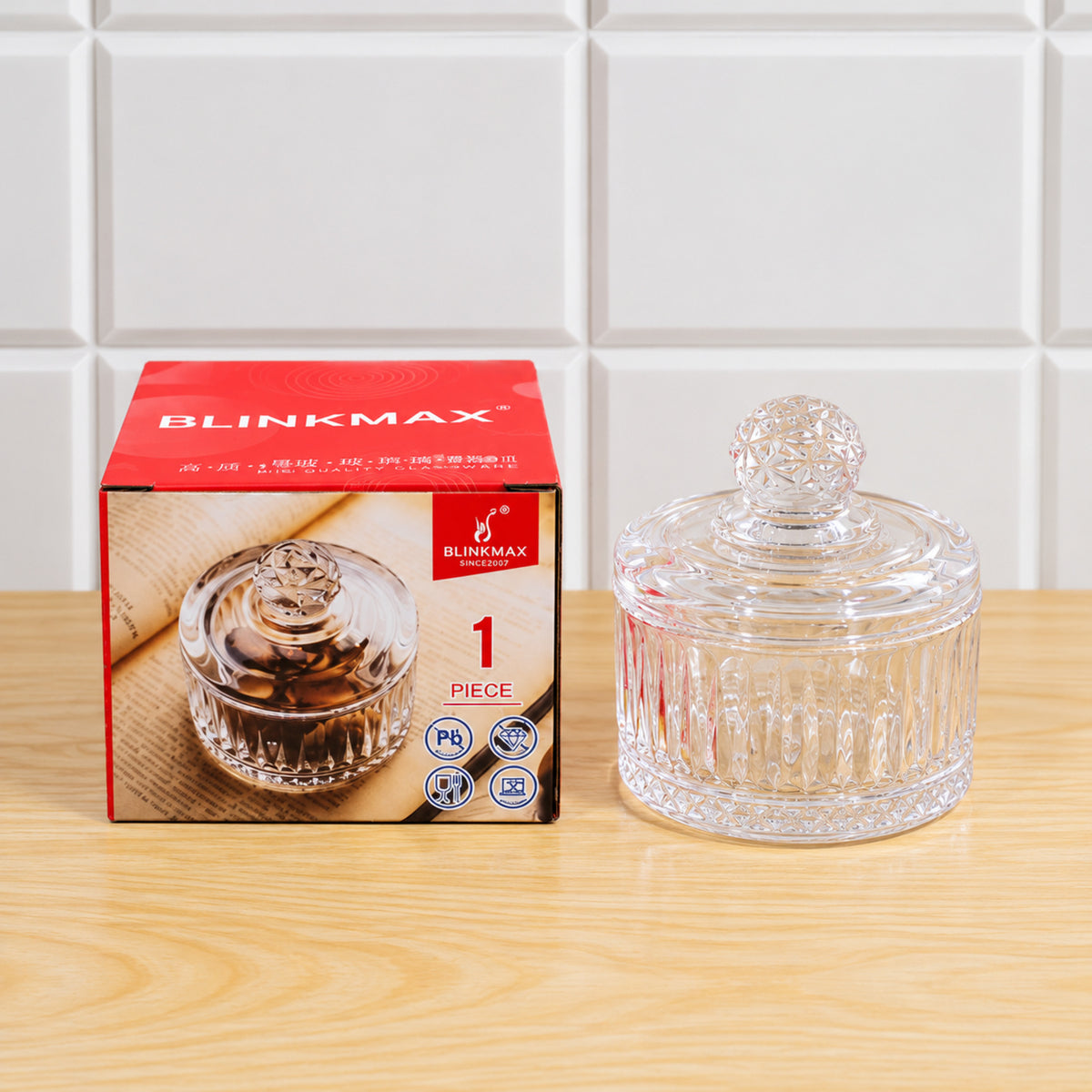 Clear glass candy bowl with lid next to its red product box, both resting on a light wooden surface.