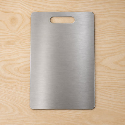 A rectangular brushed stainless steel cutting board with an integrated handle, positioned centrally on a light wooden