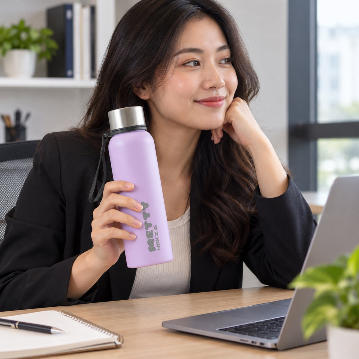 A woman holds a light purple Metty water bottle with a silver cap and strap while sitting at a desk with a laptop.