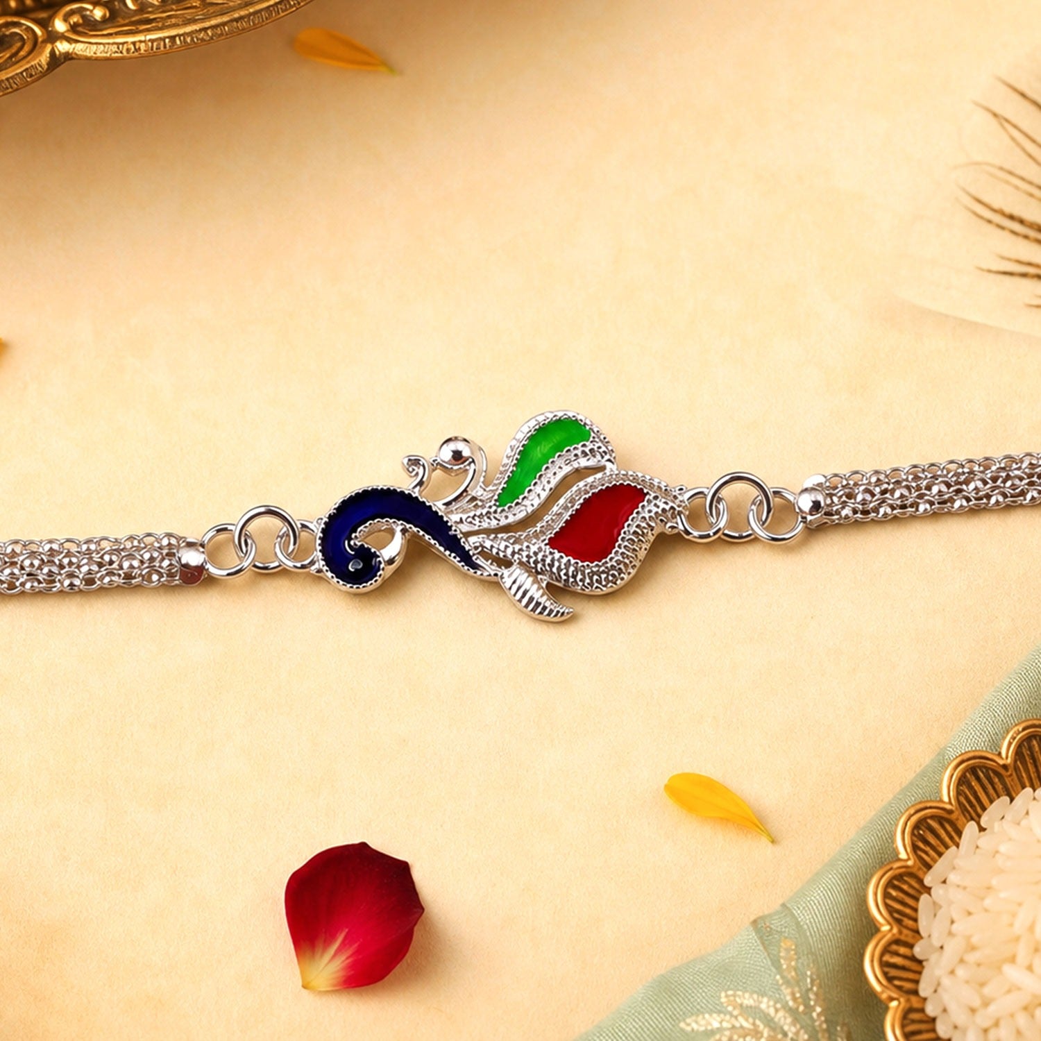 A silver chain bracelet with a central peacock motif, featuring blue, green, and red enamel accents, rests on a light yellow