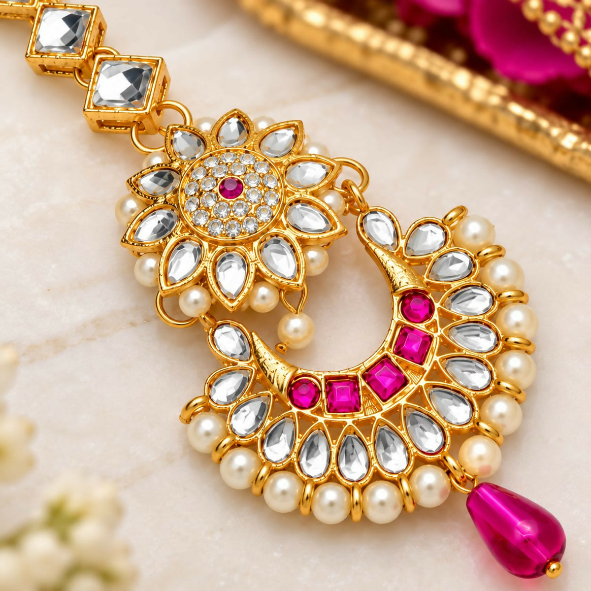 Close-up of a gold maang tikka with clear and fuchsia stones, surrounded by small white pearls, on a light marble background.