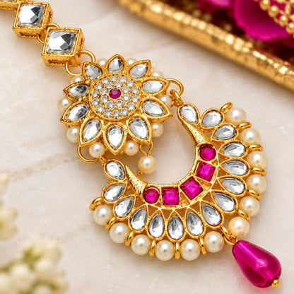 Close-up of a gold maang tikka with clear and fuchsia stones, surrounded by small white pearls, on a light marble background.