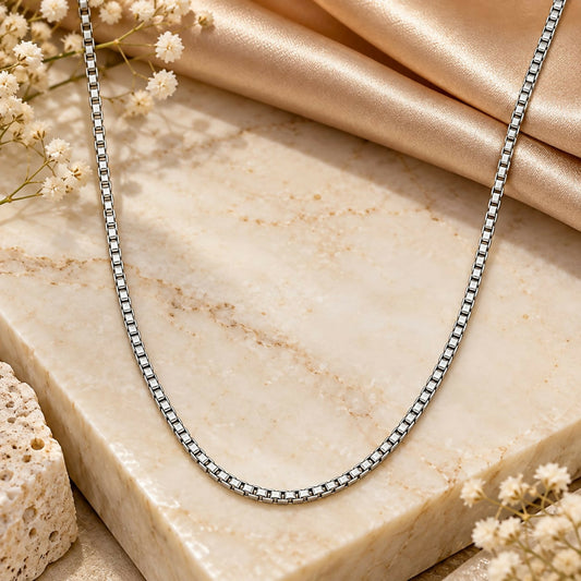 A silver-toned box link chain necklace rests on a textured stone surface with delicate white flowers.