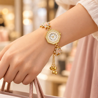 A person's wrist wears a gold-tone bracelet watch with crystal flowers and hanging charms, holding a shopping bag.