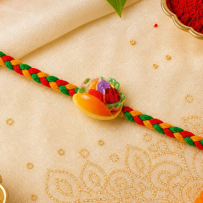 A colorful Dori Rakhi with a fruit basket charm sits on a cream-colored cloth with gold embroidery, alongside red powder in
