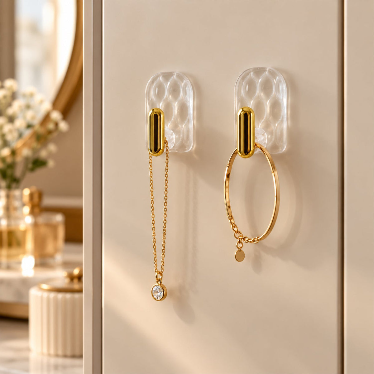 Two clear plastic hooks with gold accents hold a delicate gold chain necklace and a gold bracelet on a light wall.