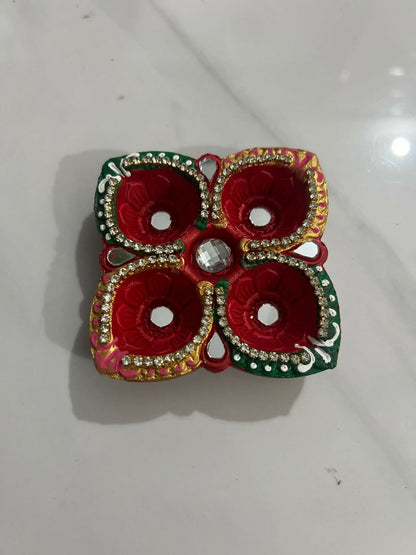A red and green four-cup diya, adorned with mirrors and small stones, rests on a light gray tiled floor.