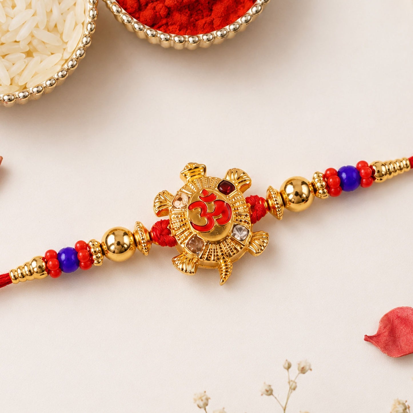 A gold tortoise charm with a red Om symbol is centered on a red thread, flanked by gold beads and red and blue smaller