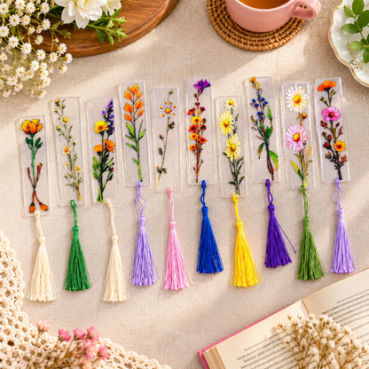 Ten transparent bookmarks with diverse floral designs and colorful tassels are arranged on a light surface.