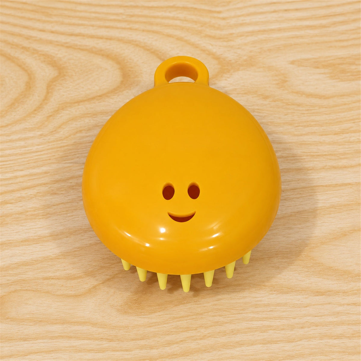 A yellow silicone scalp brush with light green bristles and a smiley face is positioned on a light wooden surface.