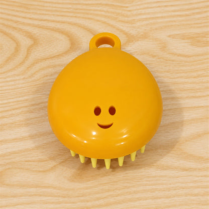 A yellow silicone scalp brush with light green bristles and a smiley face is positioned on a light wooden surface.