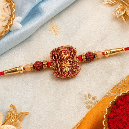 A red and gold Ganesha bracelet with rudraksha beads and golden caps, resting on a light blue and gold patterned cloth.