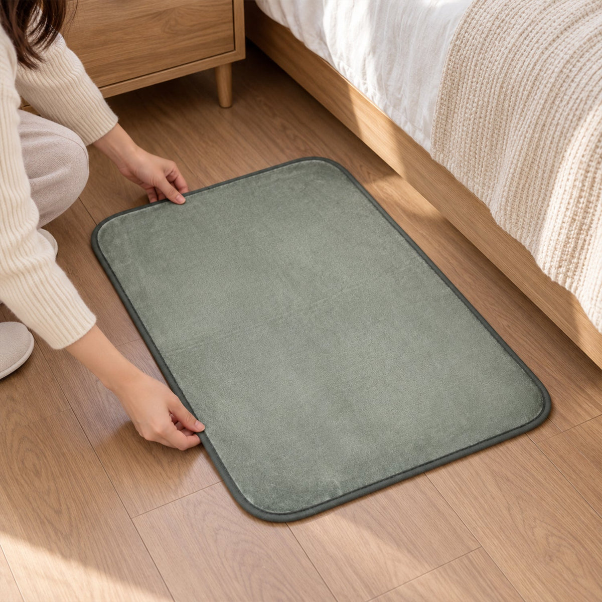 Hands adjusting a rectangular gray floor mat on a wooden floor next to a bed and nightstand.