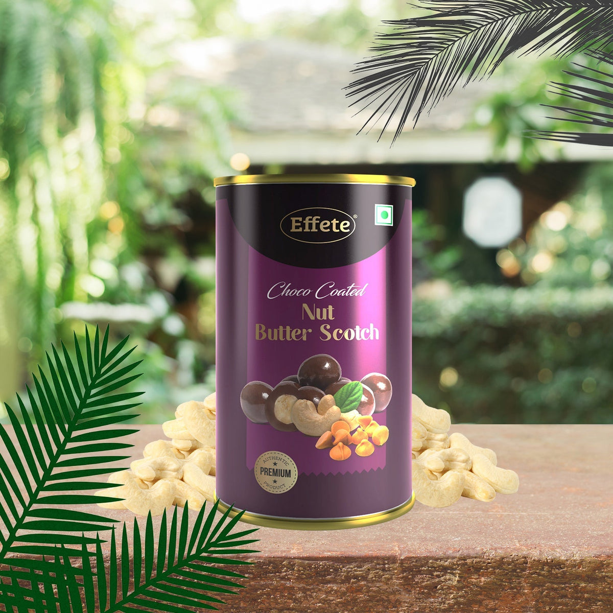 A purple and gold tin of chocolate-coated nuts sits on a wooden surface, surrounded by cashews and palm fronds.