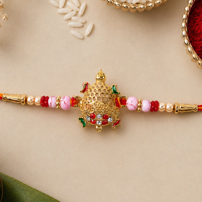 A gold turtle charm with red and white gems is centered on a red and gold beaded thread, with loose rice grains in the