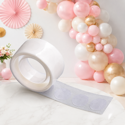A roll of clear glue dots on a white paper backing, with a pink and gold balloon arch in the background.