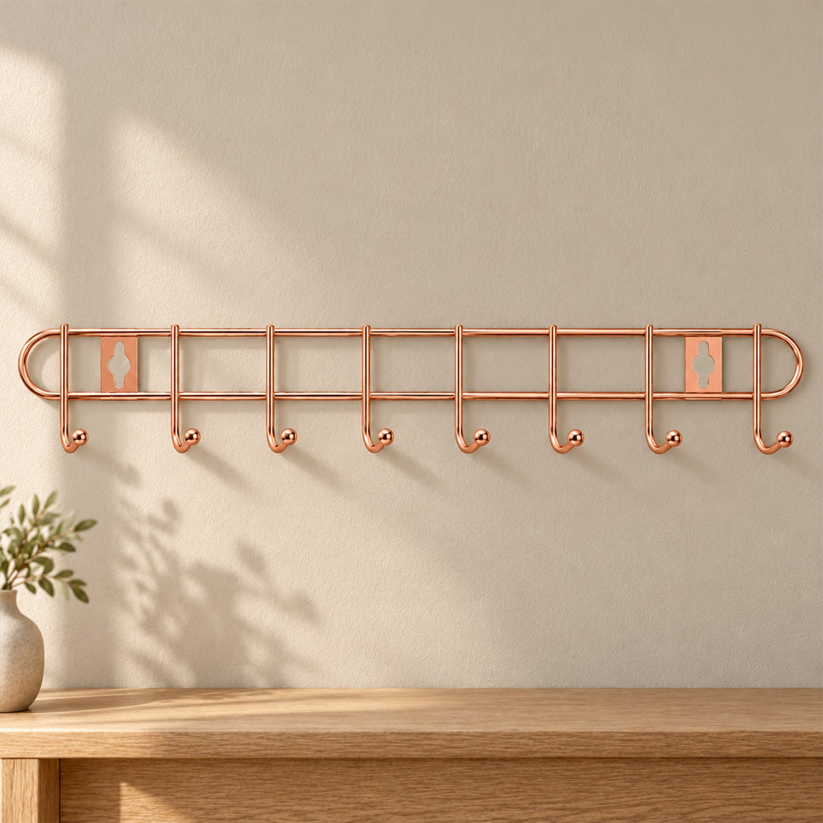 A rose gold metal wall hanger with eight hooks is mounted on a light beige wall above a wooden table.