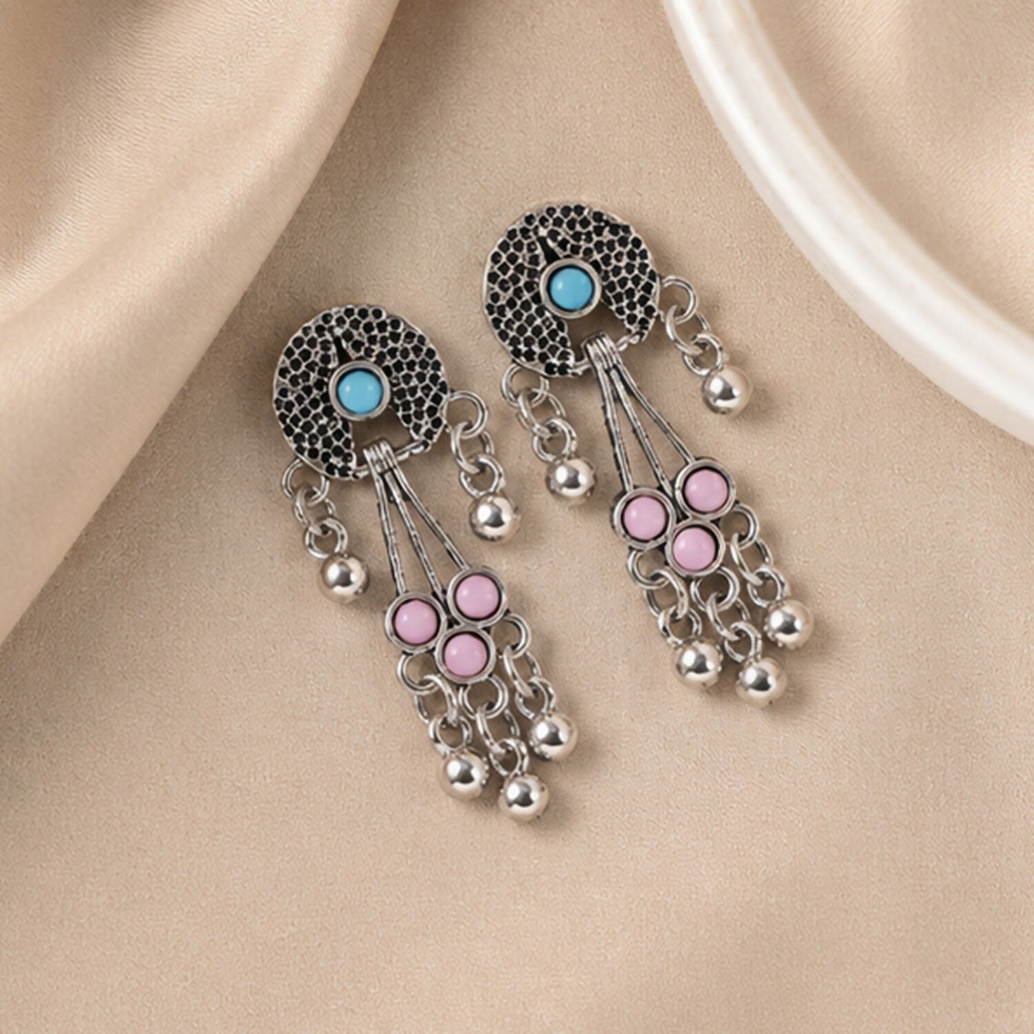 Oxidised silver long drop earrings with pink and turquoise stones and ghungroo bells lie on a light brown fabric surface.