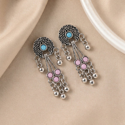 Oxidised silver long drop earrings with pink and turquoise stones and ghungroo bells lie on a light brown fabric surface.