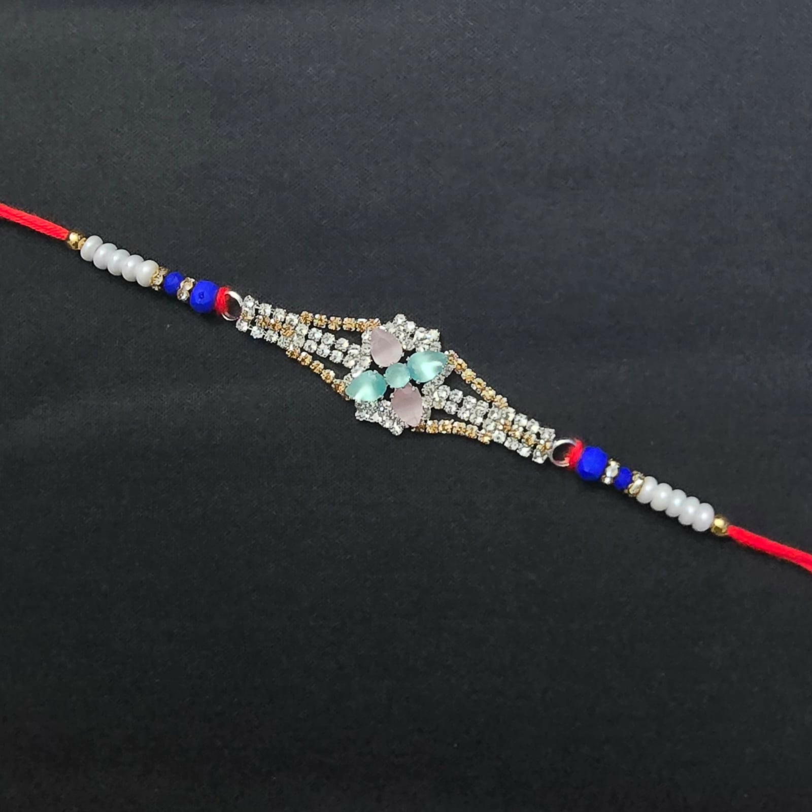 A red thread bracelet with a central diamond-studded floral design featuring pink and blue-green crystals, on a plain black