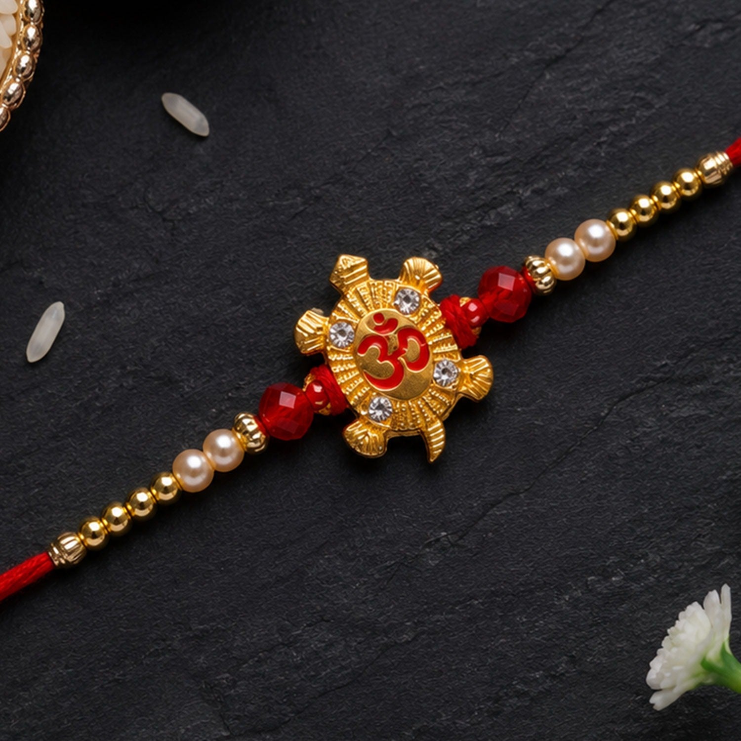 A red thread bracelet with a gold turtle charm, red beads, and white pearls on a dark background with scattered rice grains.
