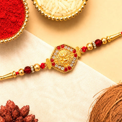 A golden Ganpati motif with red and clear stones is centered on a red thread with golden beads, next to bowls of rice and