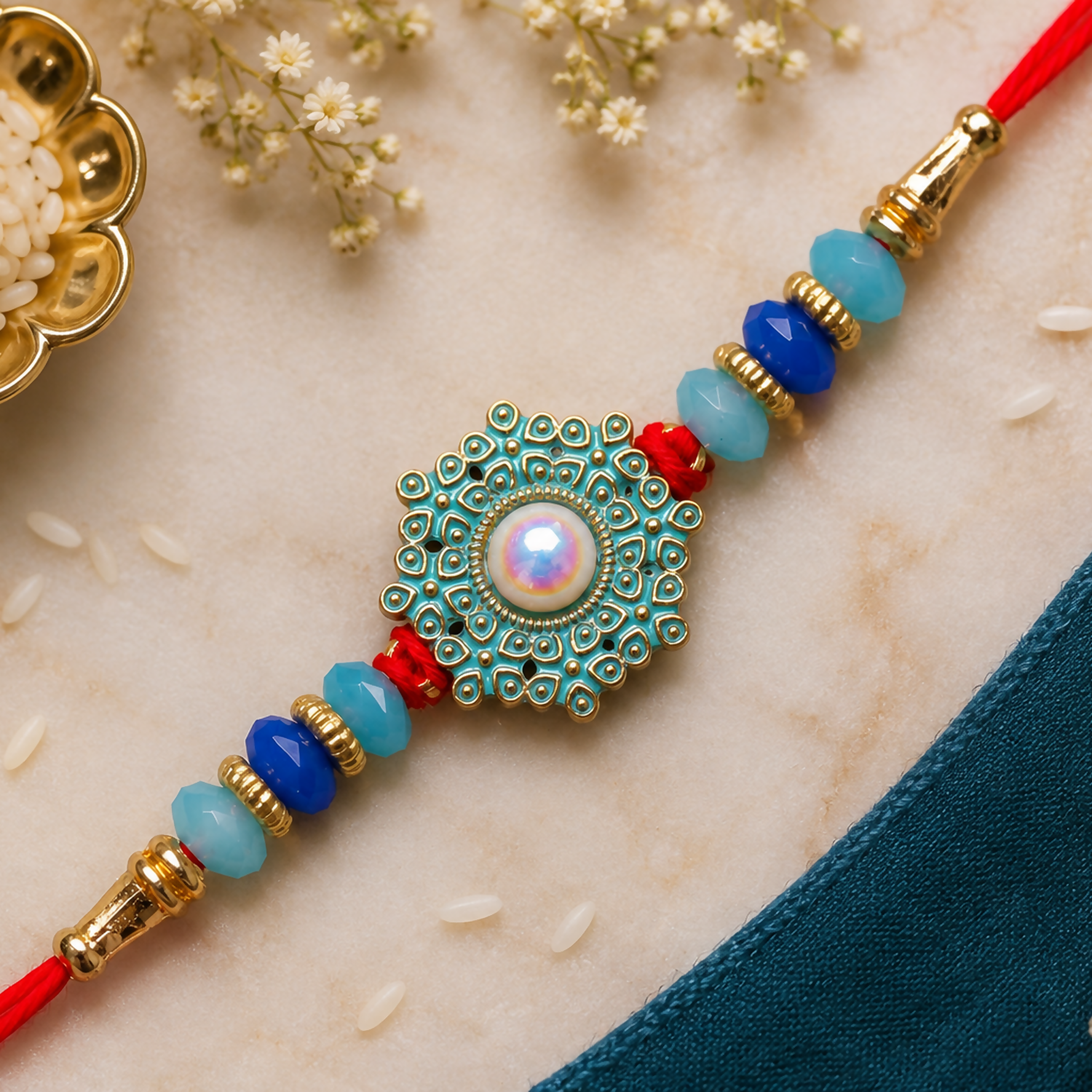 A red thread with a teal and gold medallion, featuring a central pearl, is adorned with blue and light blue beads.