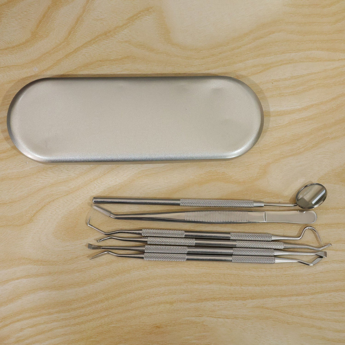 The closed metal dental tool case and the six individual stainless steel tools are neatly arranged on a light wooden surface.