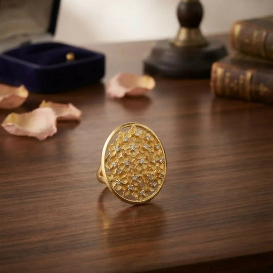 A gold-tone ring with an oval floral cluster design and small sparkling stones rests on a wooden table.