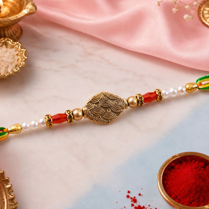 A Rakhi with a central antique gold oval charm, flanked by red crystal and white pearl beads, on a pink and white background