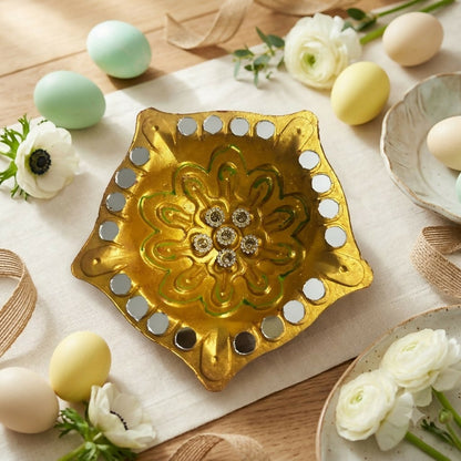 A gold star-shaped diya with a floral center and mirror border sits on a white cloth with pastel eggs and flowers.