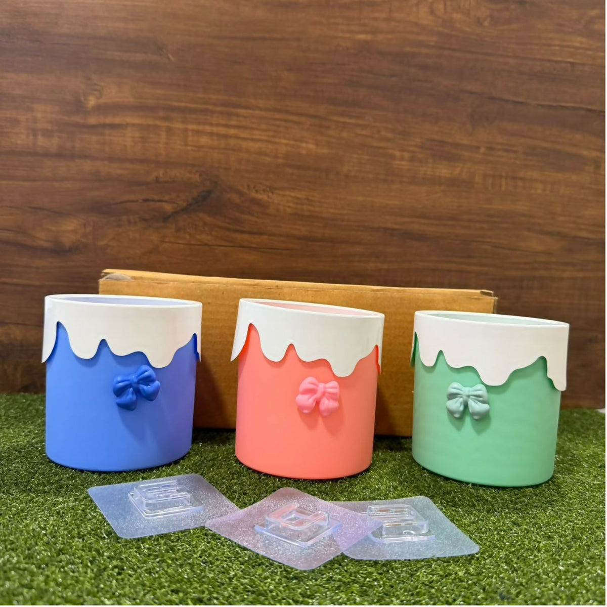 Three empty pastel organizers with adhesive hooks on artificial grass against a wooden background.
