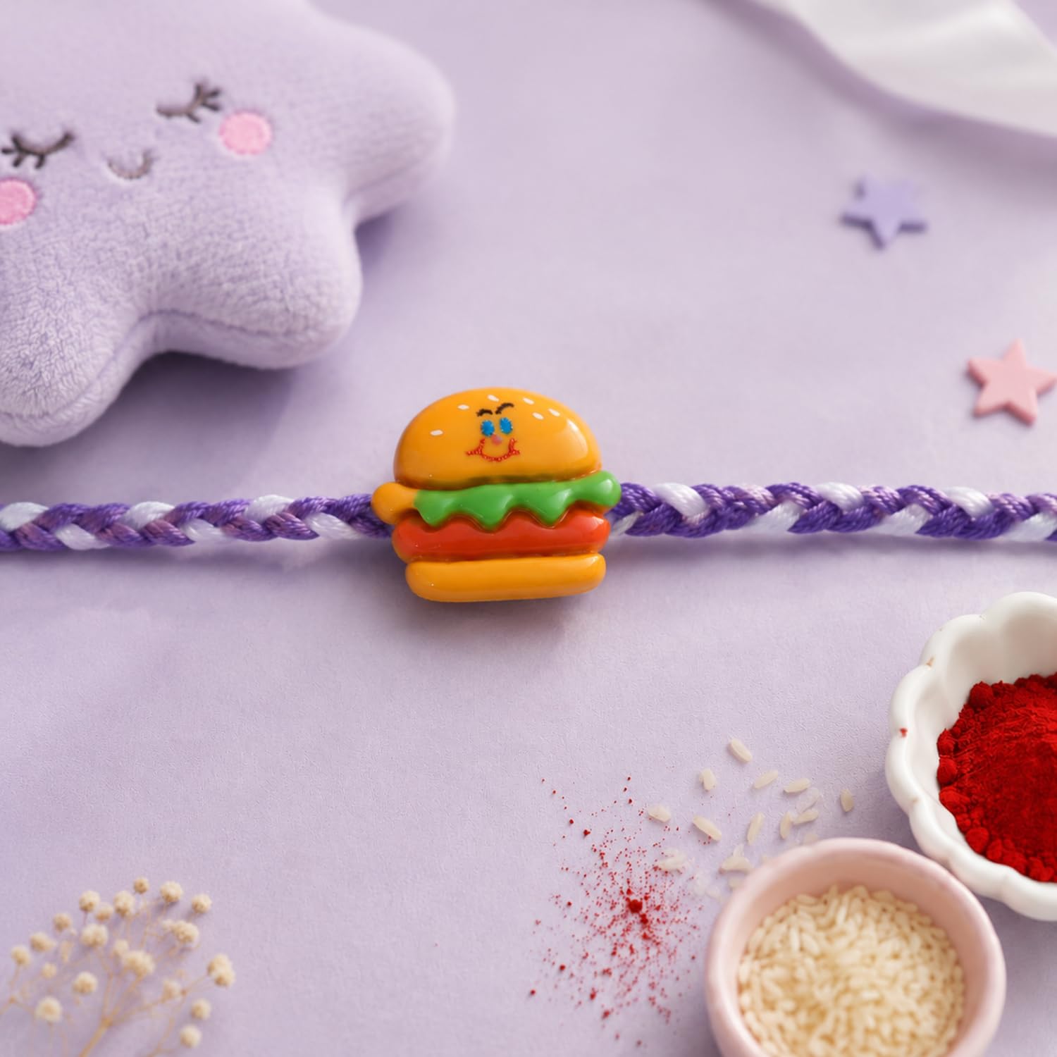 A purple and white braided thread features a small, colorful hamburger charm, with a plush star and rice grains scattered