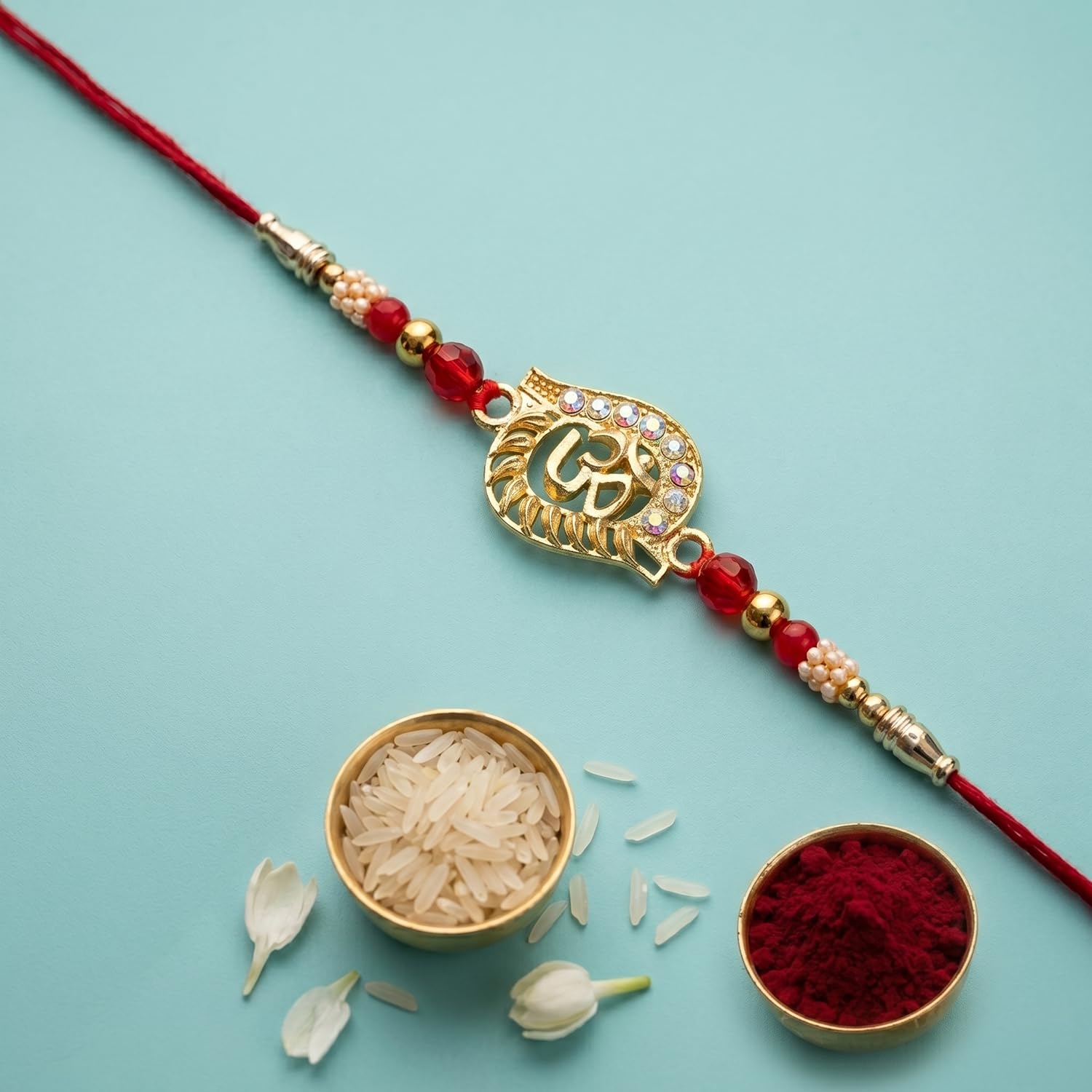 A red thread bracelet with a gold Om symbol charm and red and white beads is laid out next to small bowls of rice and red
