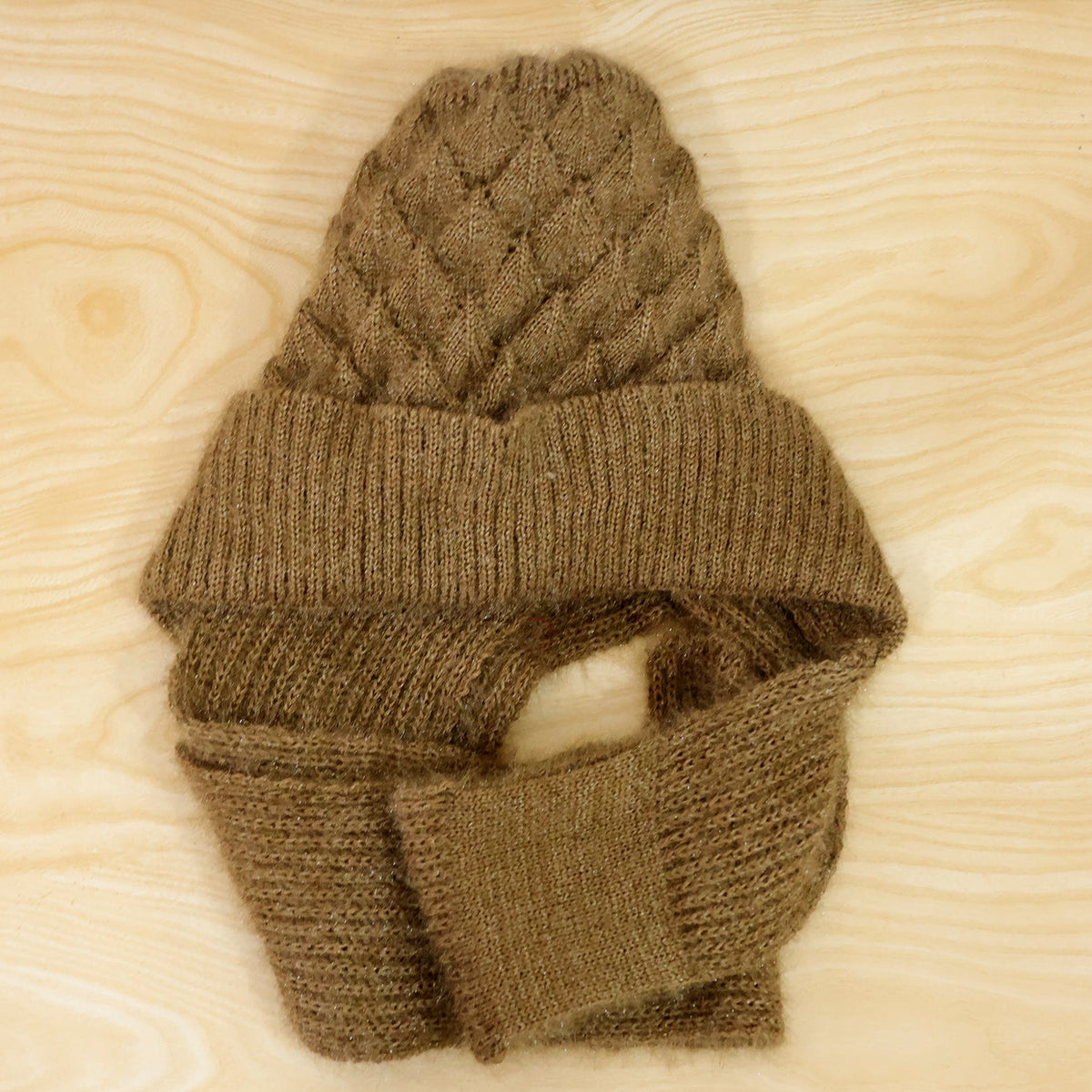 A brown knitted cap with an attached scarf is laid flat on a light wood surface, showcasing its texture.