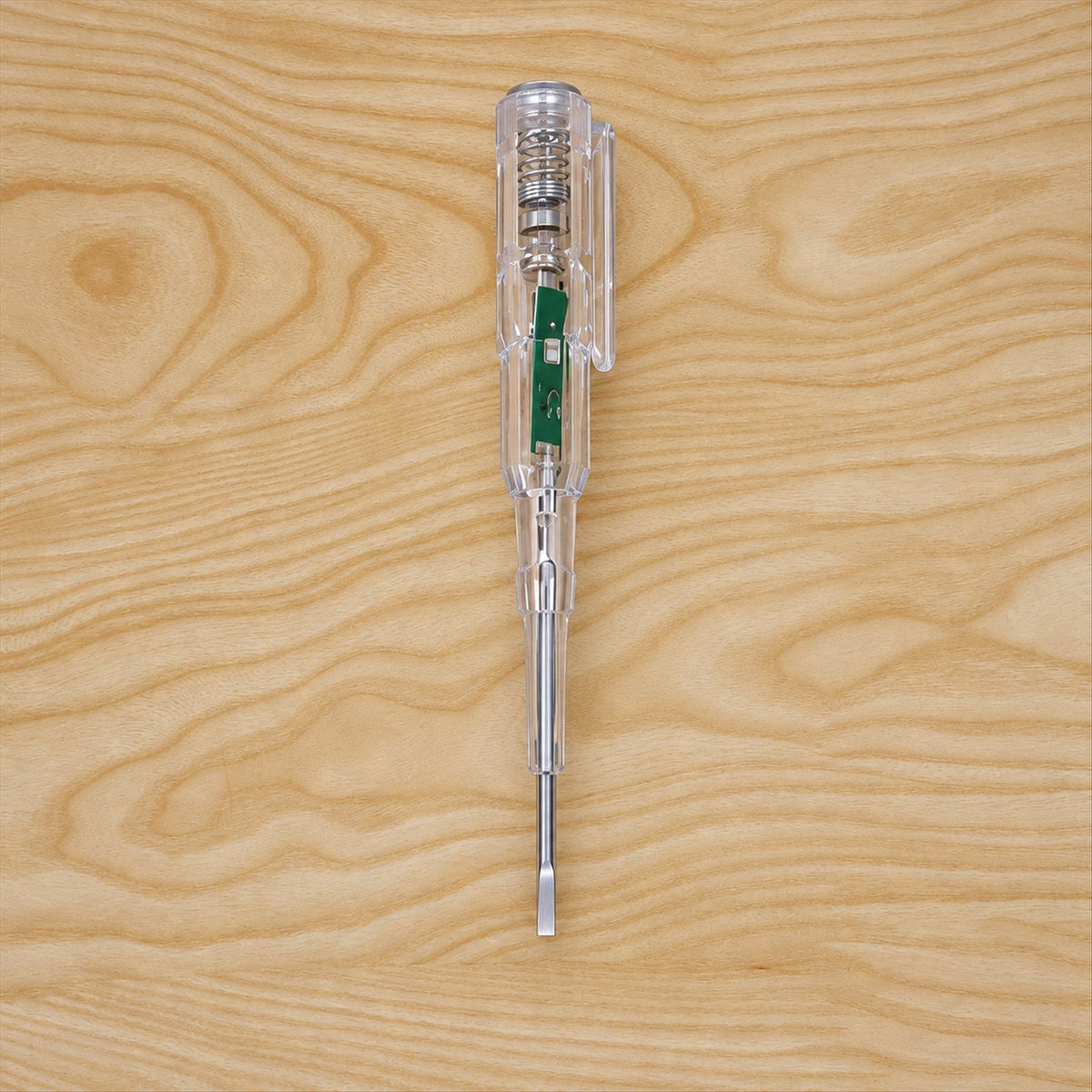 A transparent electrical tester screwdriver with a flat head and internal green circuit board is centered on a light wooden