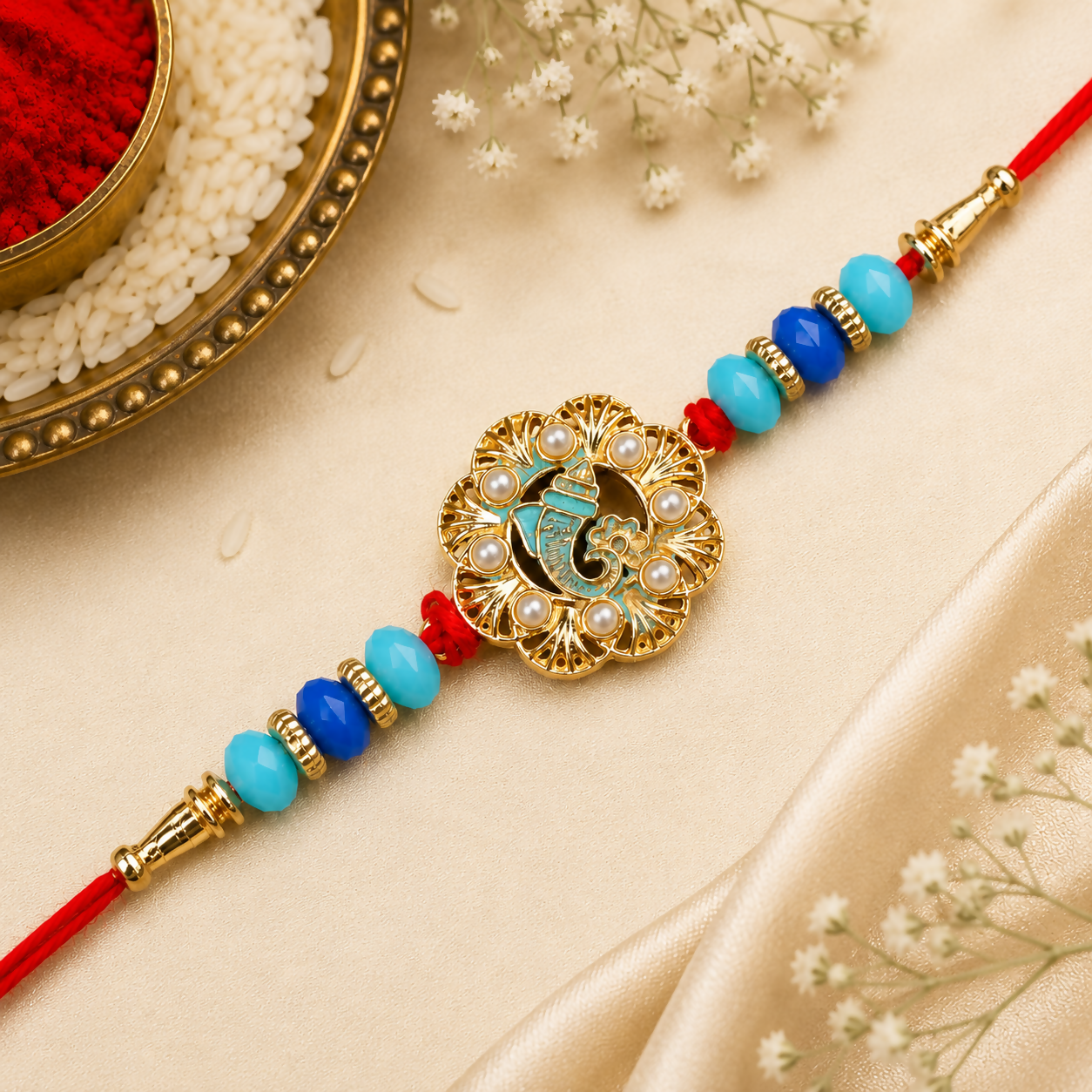 The ornate gold Ganesha charm, featuring blue enamel and small pearls, is flanked by blue and turquoise beads on a red
