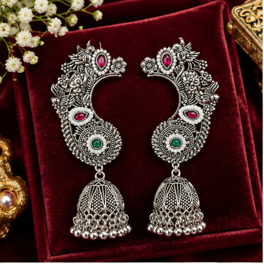 A pair of ornate oxidized silver earrings with red and green stones, white enamel, and bell drops on a red velvet surface.
