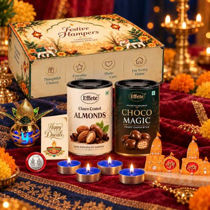 A Diwali gift set with a decorative box, two chocolate tins, a kalash, lit tealights, and a greeting card on a red cloth.