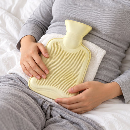 A person lies on a white bed, holding a light yellow hot water bottle wrapped in a white towel on their abdomen.