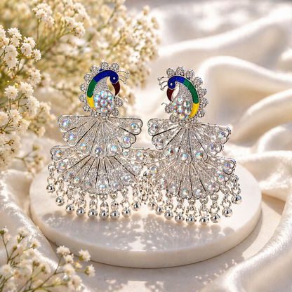 A pair of silver peacock earrings with colorful enamel and clear stones, displayed on a white surface with flowers.