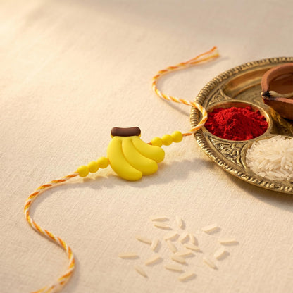 A yellow and orange bracelet with a banana charm lies on a white surface next to a brass plate holding a red powder and