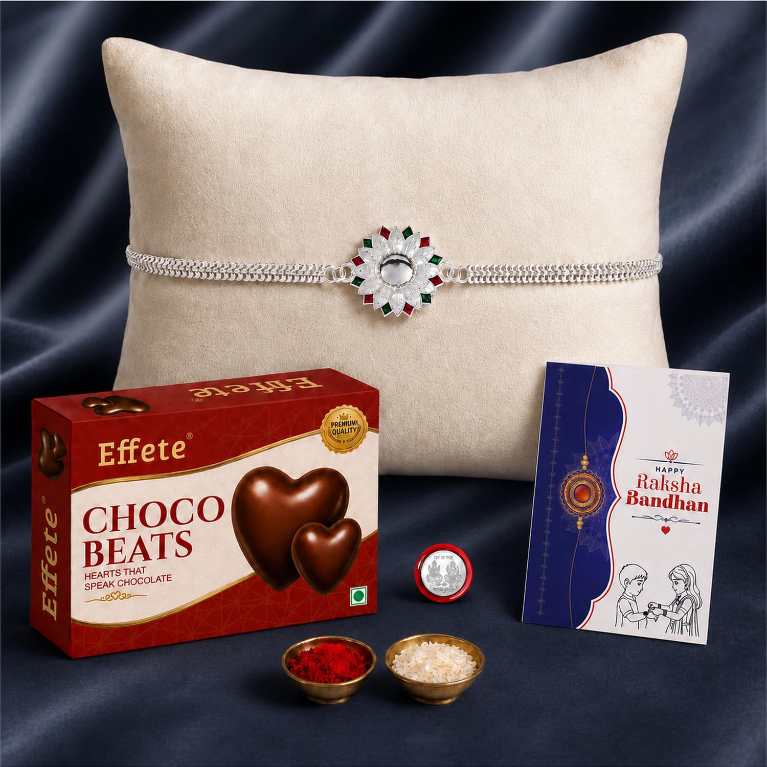 A silver-colored Rakhi with a white, red, and green floral centerpiece rests on a cream pillow, with chocolates, kumkum,