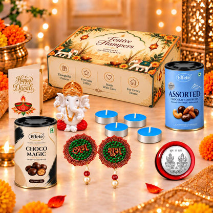 A festive gift box with a Ganesha idol, tealights, decorative hangings, a silver coin, two chocolate tins, and a greeting